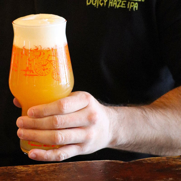 Man holding a glass of Boneshaker Juicy Haze IPA beer with 'Boneshaker Juicy Haze IPA' shirt in the background