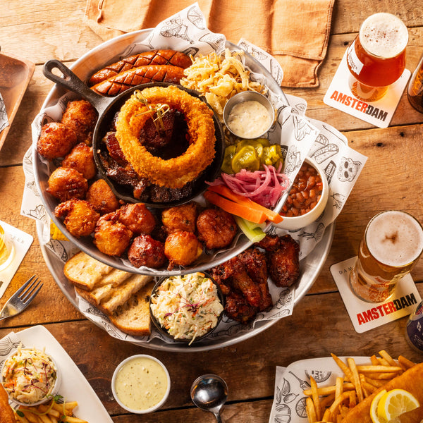 Assorted food platter with fried onion rings, wings, and sides on a wooden table with Amsterdam craft beer and snacks.