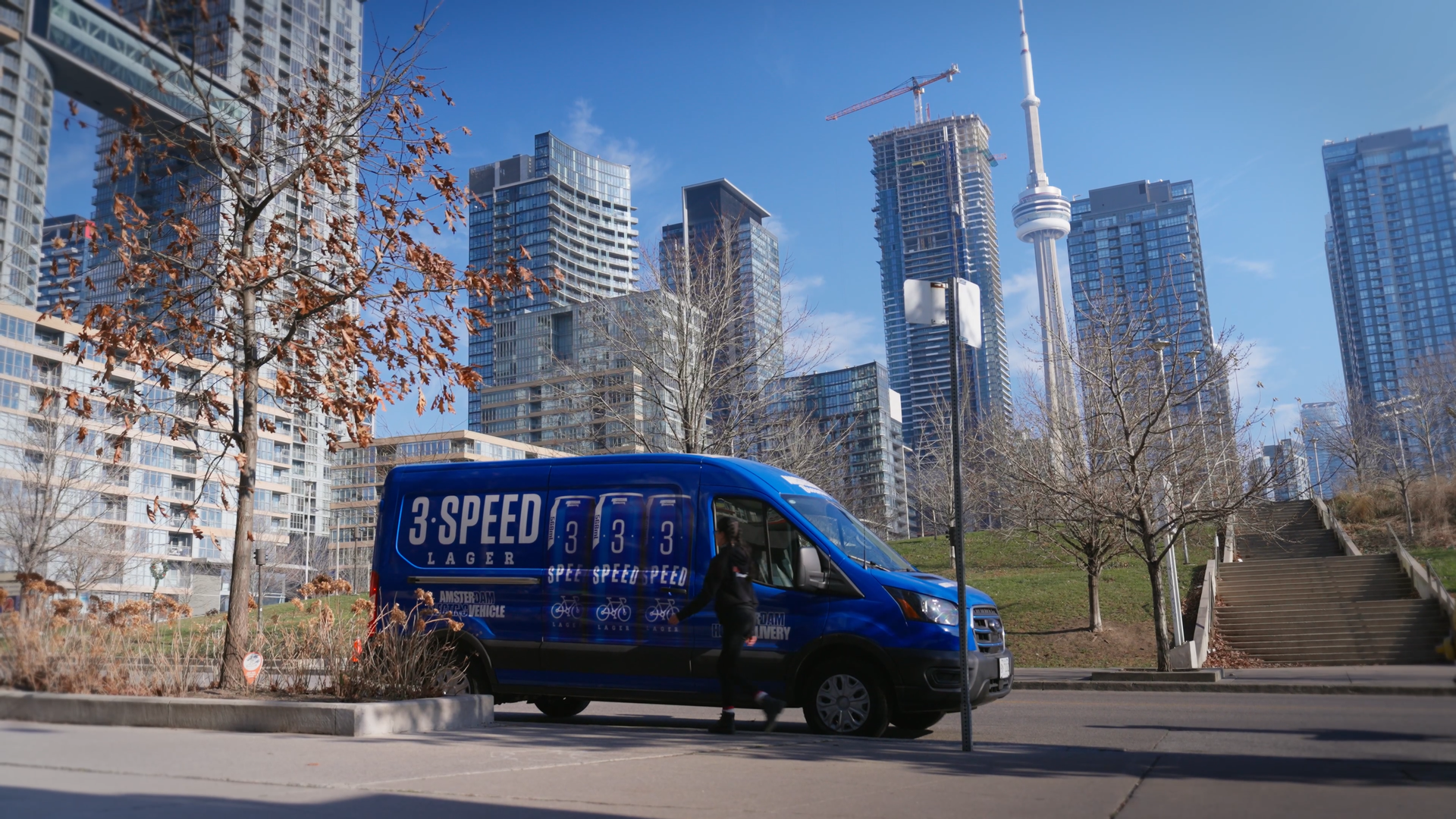 Blue van with '3 Speed Lager' branding in Toronto's urban setting with tall buildings and the CN Tower in the background on a fall morning.