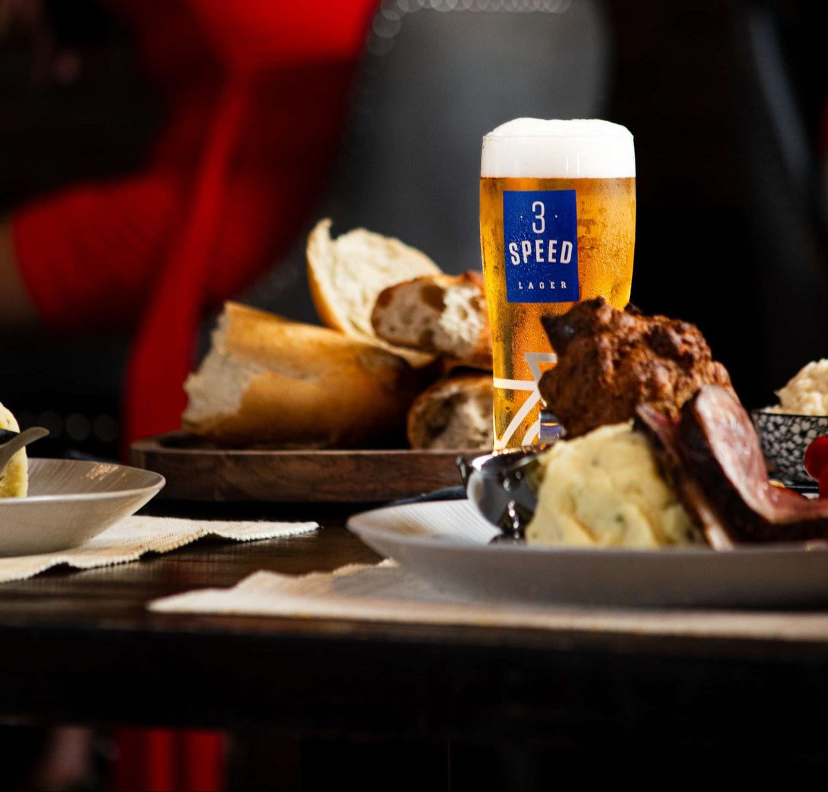 Glass of beer with '3 Speed' label on a table with food and bread.